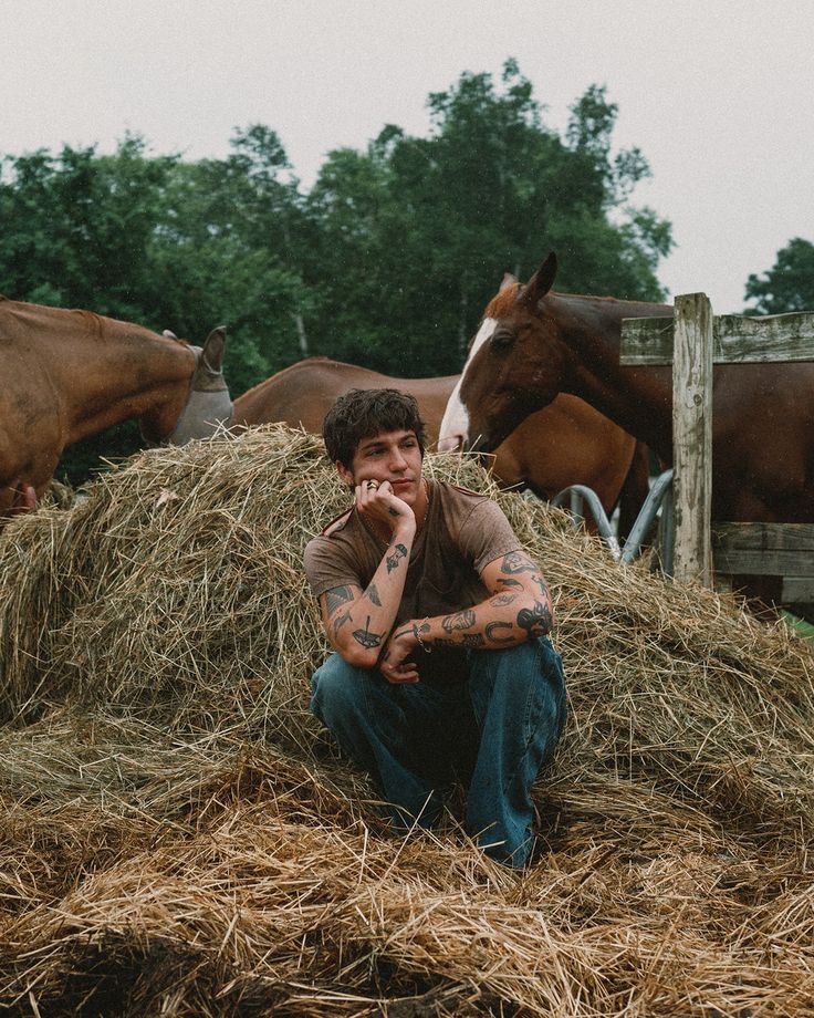 a man sitting on top of a pile of hay with horses in the back ground