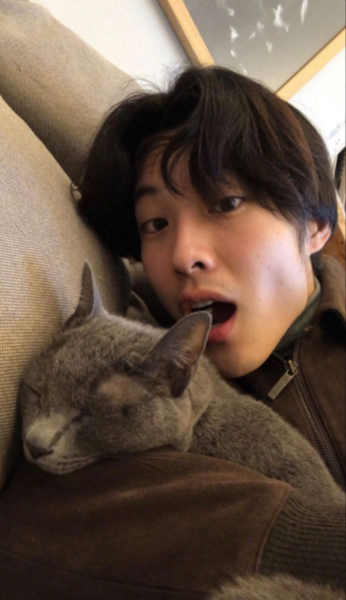 a young man is cuddling with a cat on his lap and making a funny face