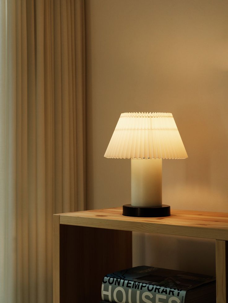 a table lamp sitting on top of a wooden table next to a book and window