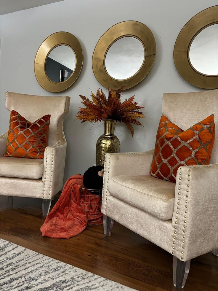 two white chairs with orange pillows in front of three gold mirrors on the wall above them