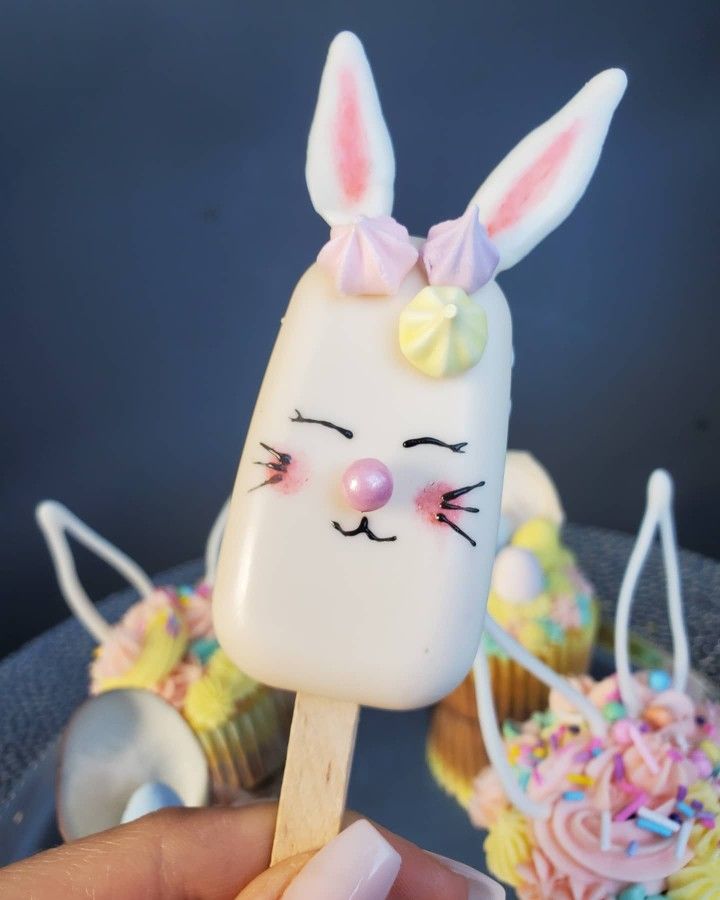 a hand holding a popsicle with an image of a bunny on it and cupcakes in the background