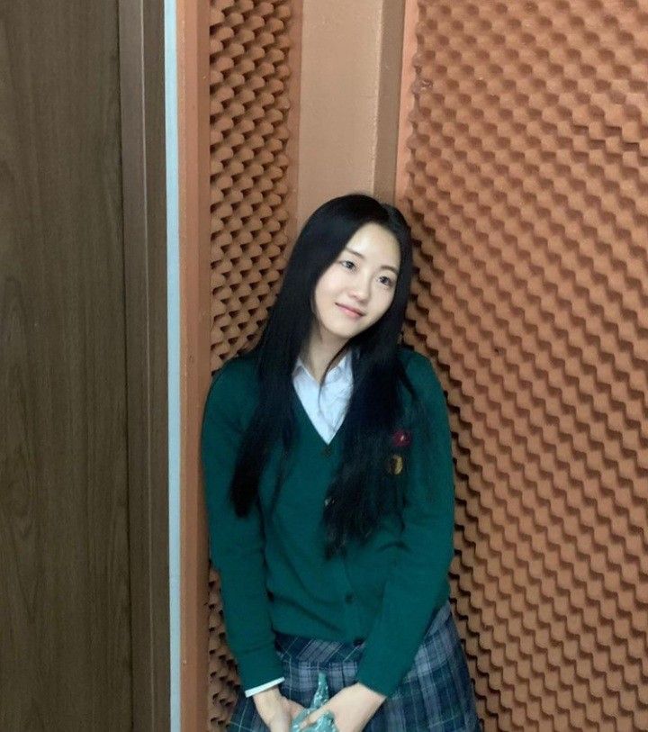 a woman in a green sweater and plaid skirt leaning against a wall