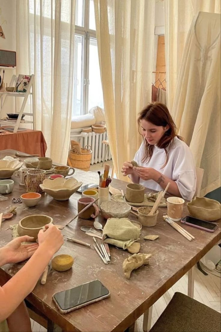 two women are sitting at a table making bowls and spoons with pottery on it