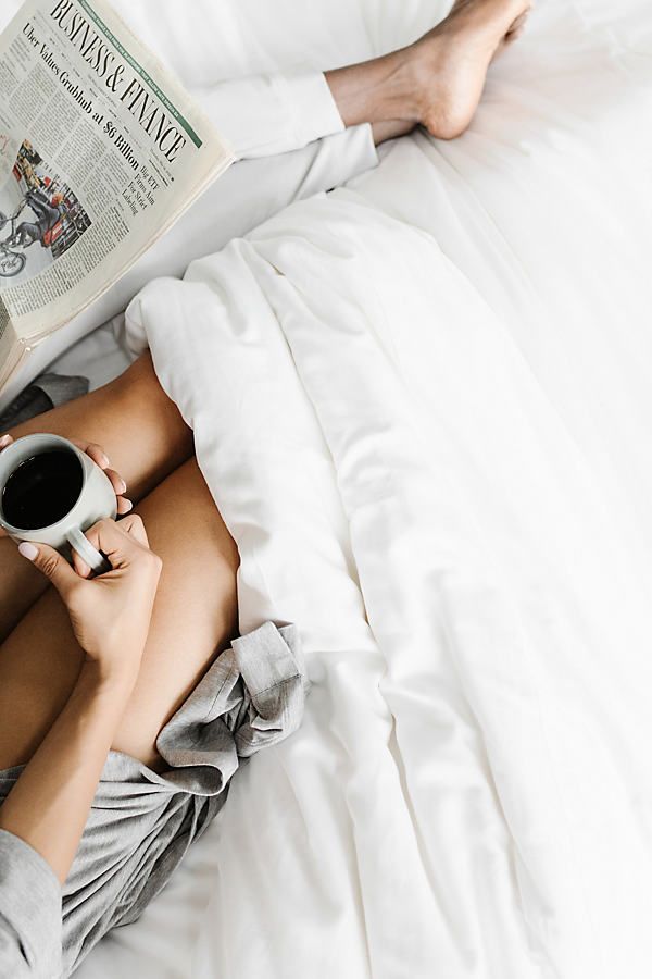 a person laying in bed reading a book and holding a cup of coffee with both hands