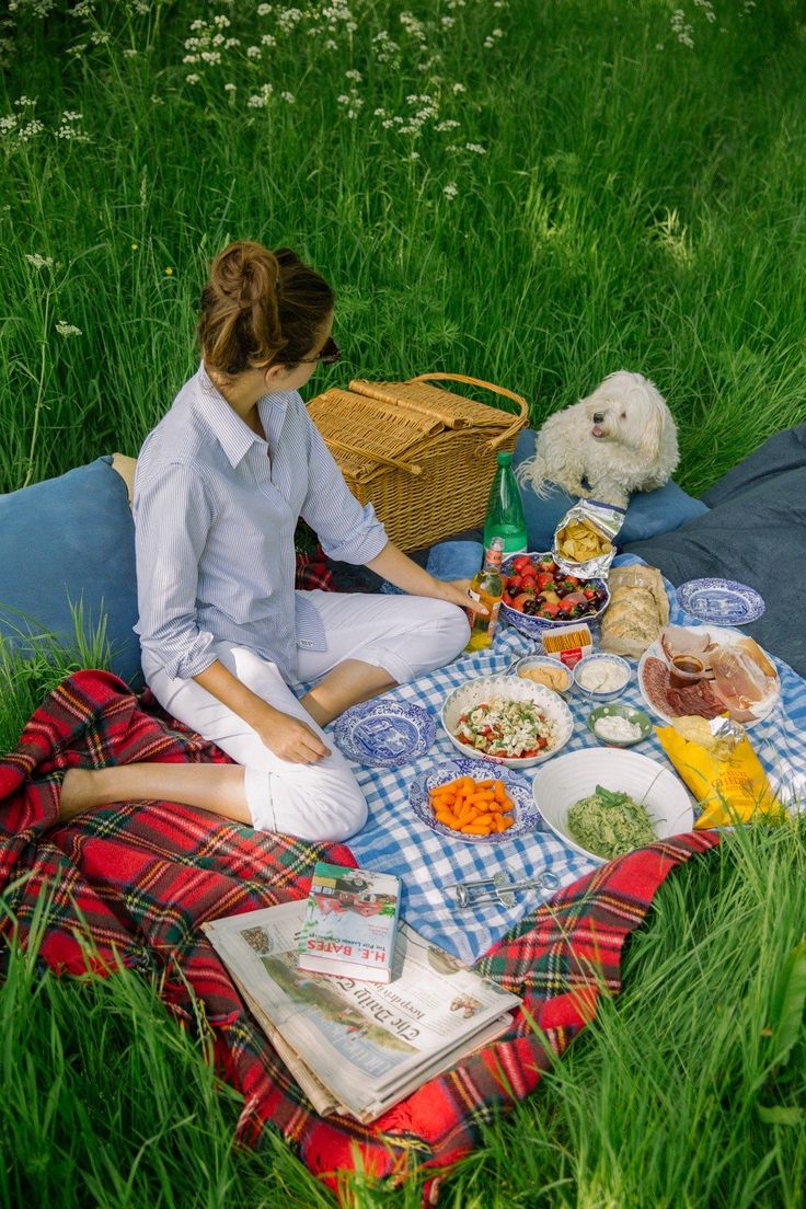 a woman sitting in the grass with food on her picnic blanket next to a dog