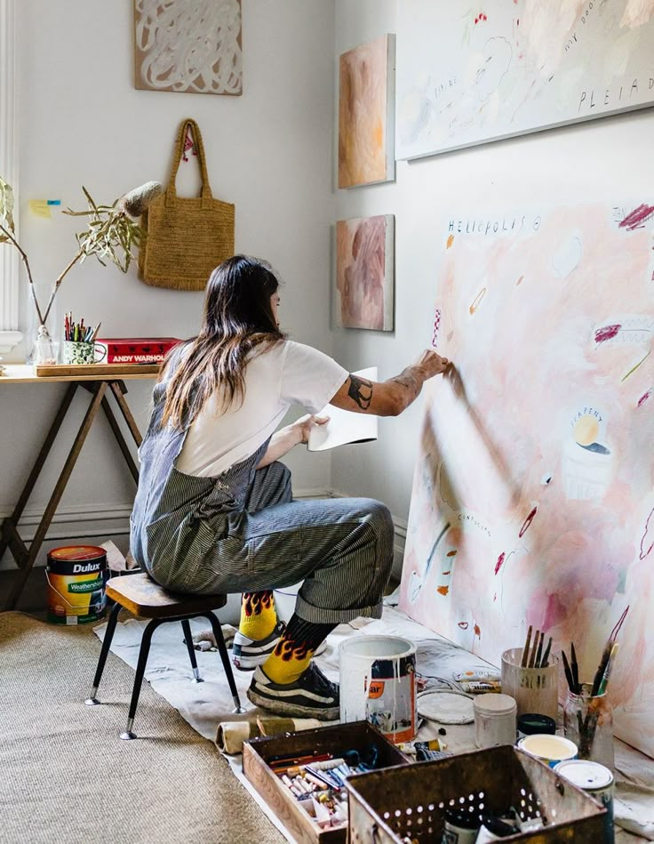 a woman sitting on a chair in front of a wall covered with paintings and art supplies