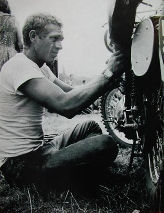 a man sitting on the ground working on a motorcycle tire while another man looks on