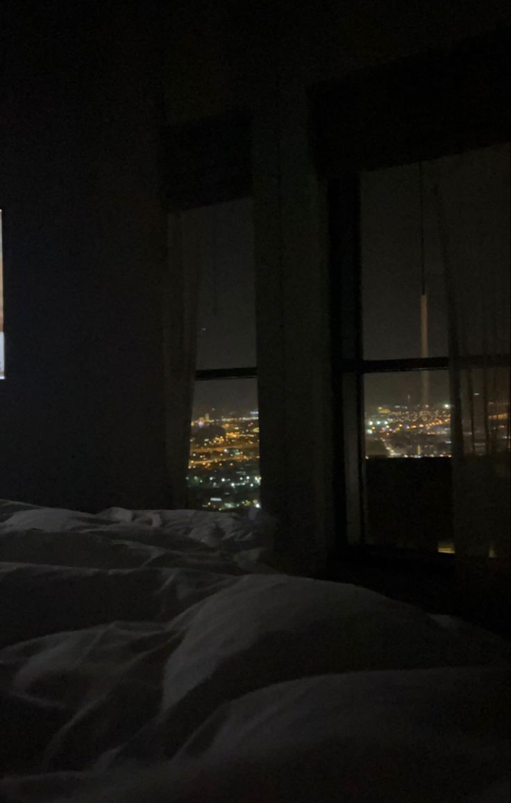 a bed in a dark room with the city lights lit up at night from behind it