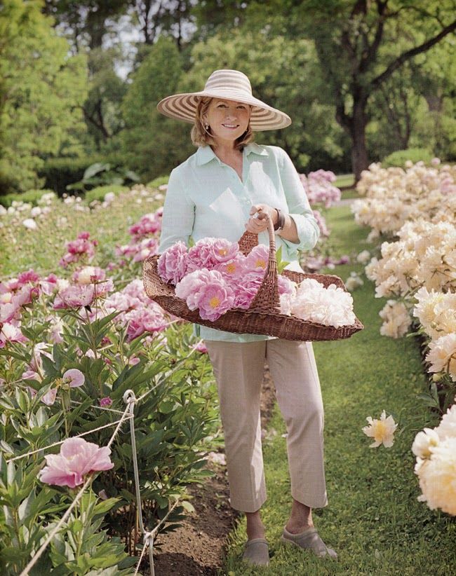 a woman holding a basket full of pink flowers in a flower garden with lots of white and pink peonies