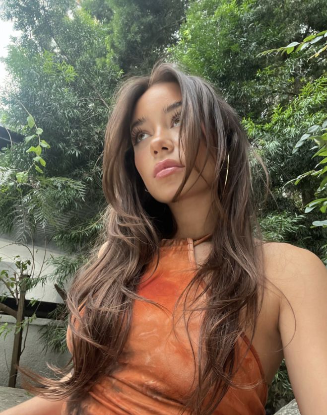 a woman with long brown hair sitting on a bench in front of trees and bushes