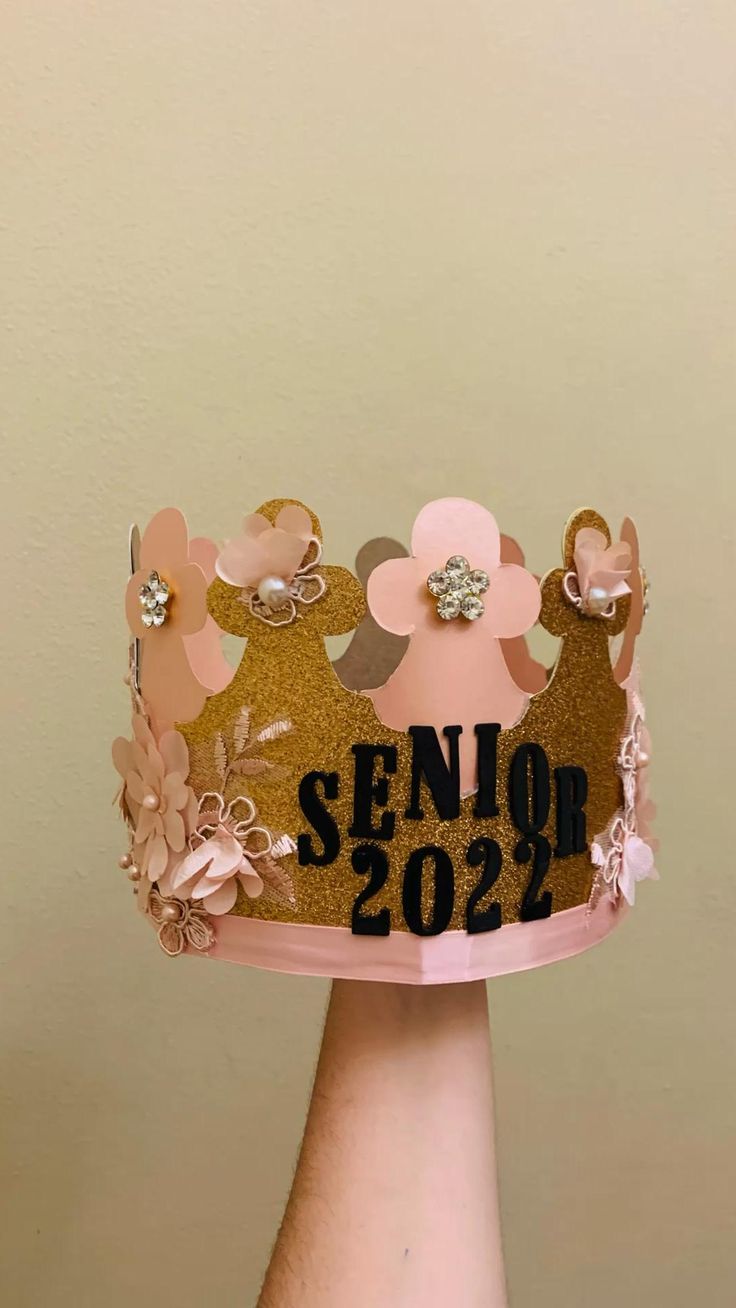 a hand holding a pink and gold crown with the words senior so2 on it