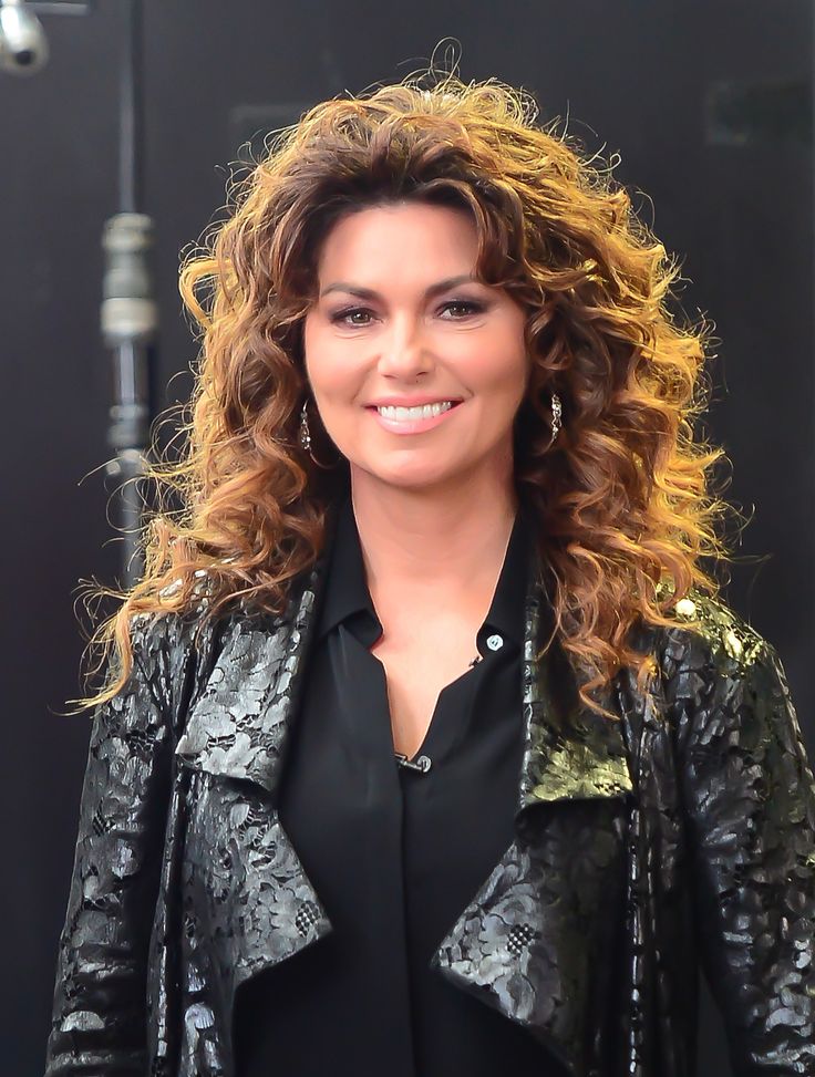 a woman with long curly hair wearing a black shirt and leather jacket smiling at the camera