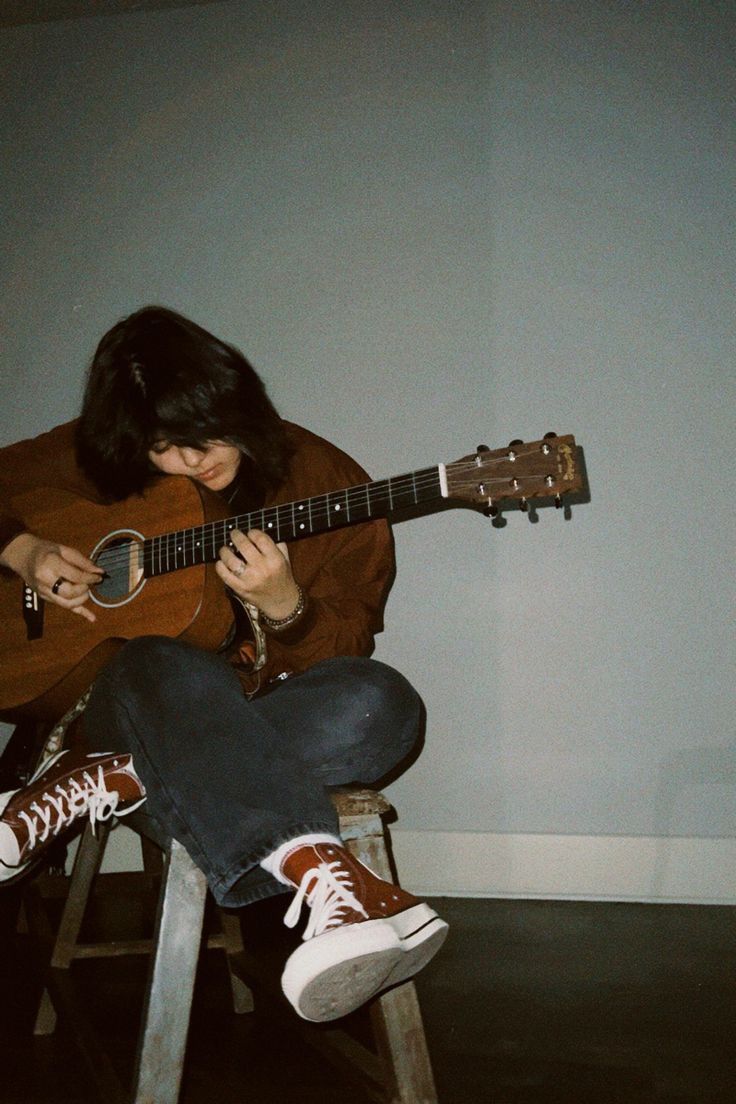 a woman sitting on a stool playing an acoustic guitar with her legs crossed and head down