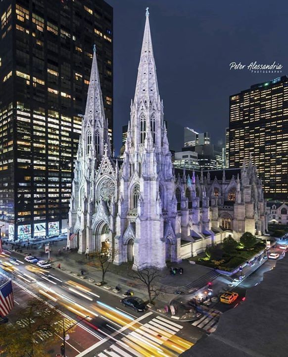 the cathedral is lit up at night in front of tall buildings and skyscrapers with cars passing by