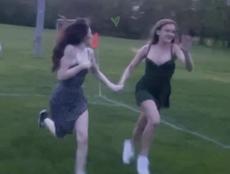 two young women running across a field holding hands and smiling at the camera, both wearing short dresses