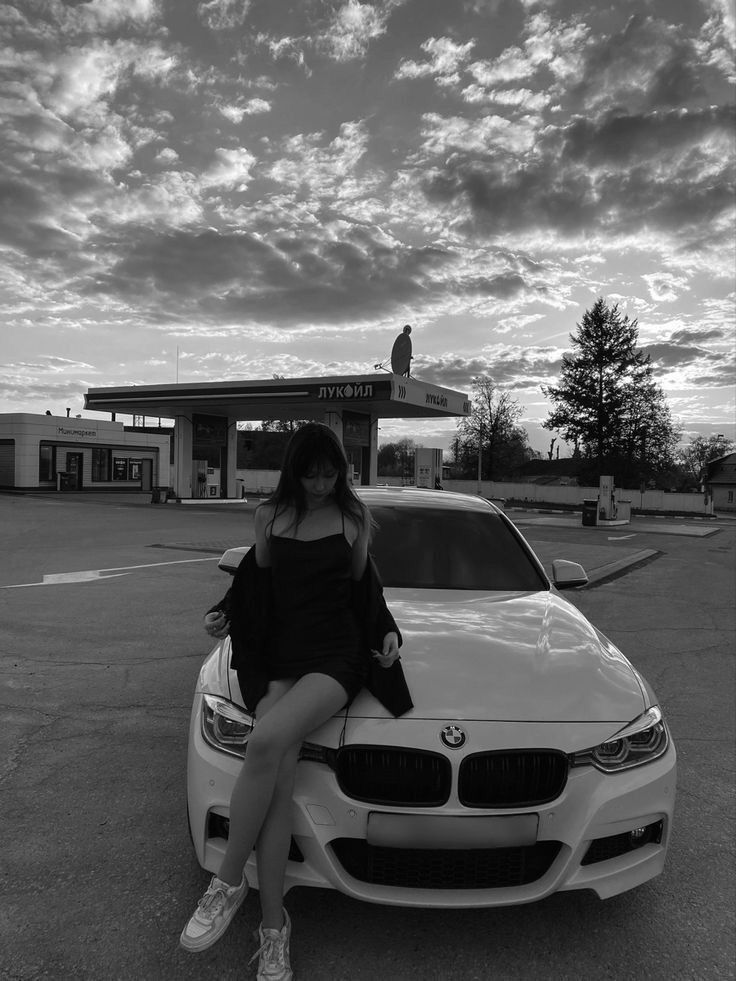 a woman sitting on the hood of a white car in front of a gas station