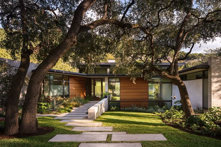 a modern house with trees and grass in the front yard, surrounded by stone walkways