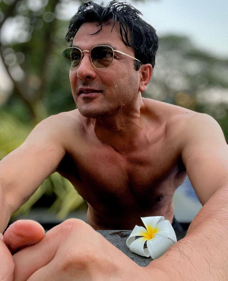 a shirtless man wearing sunglasses and holding a flower in his left hand while sitting on the beach