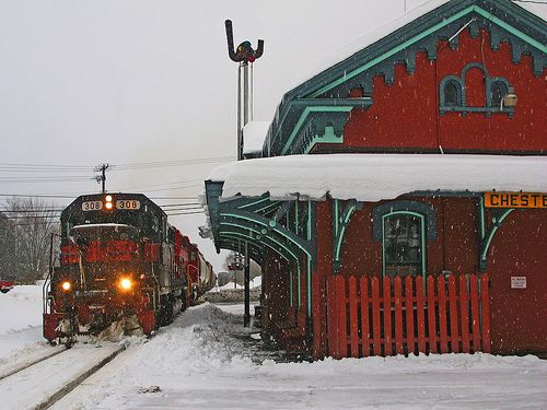 two trains on the tracks in front of a red building with green trim and snow