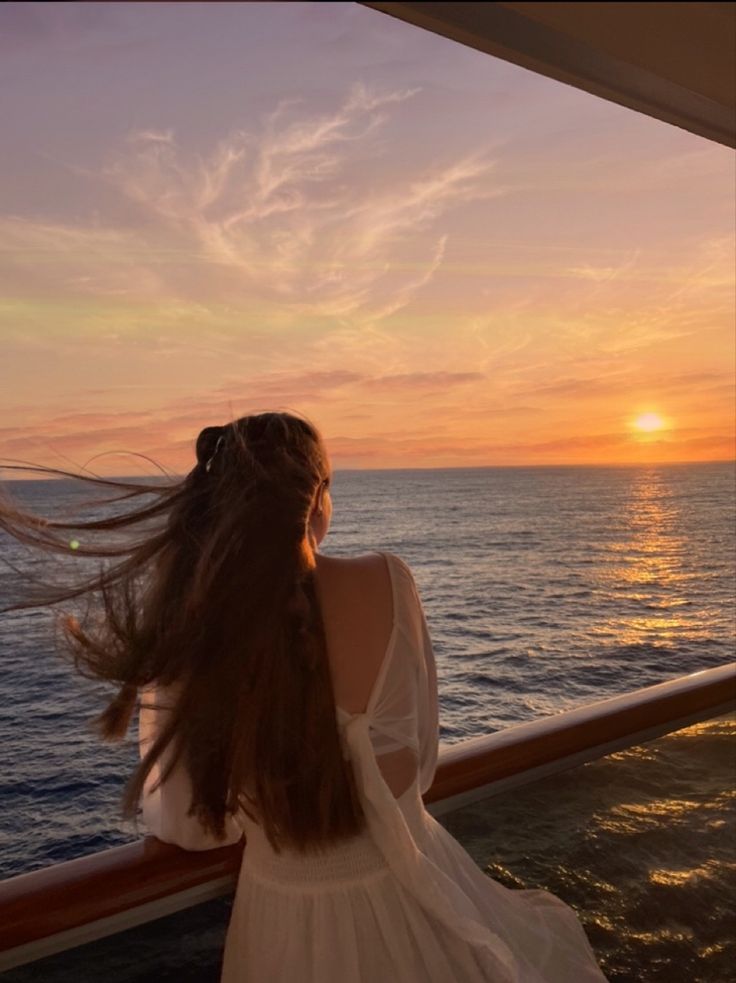 a woman standing on the deck of a boat looking out at the ocean as the sun sets