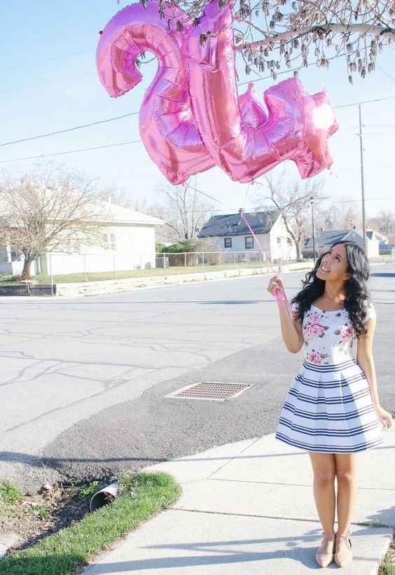 a woman in a dress and hat holding up a pink number 25 balloon while standing on the sidewalk