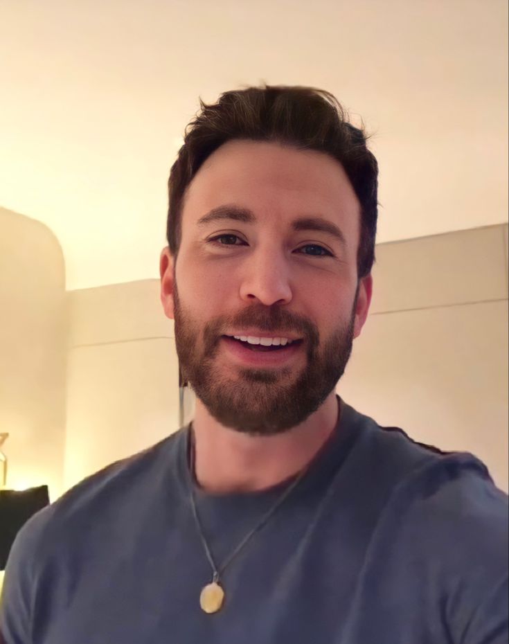 a man with a beard smiling and wearing a blue t - shirt in a living room