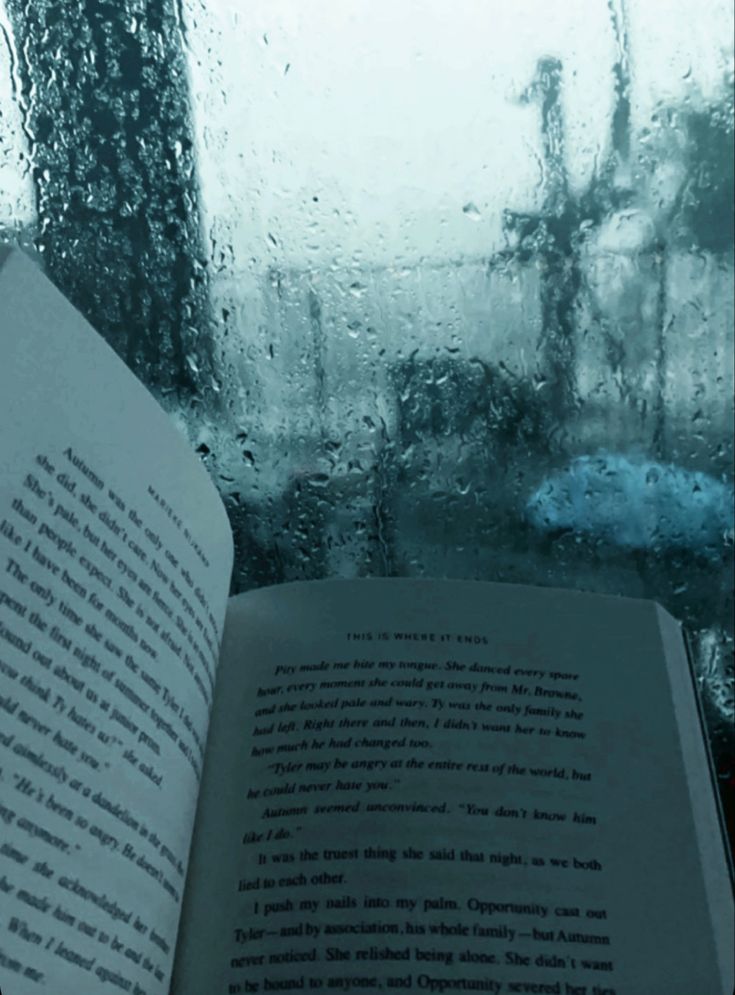 an open book sitting on top of a window sill next to rain covered windows