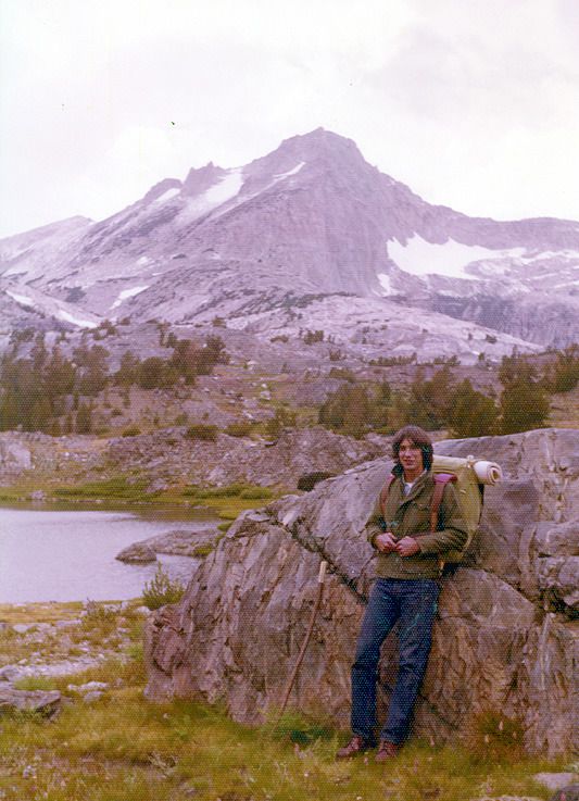 a man standing on top of a large rock next to a body of water with mountains in the background