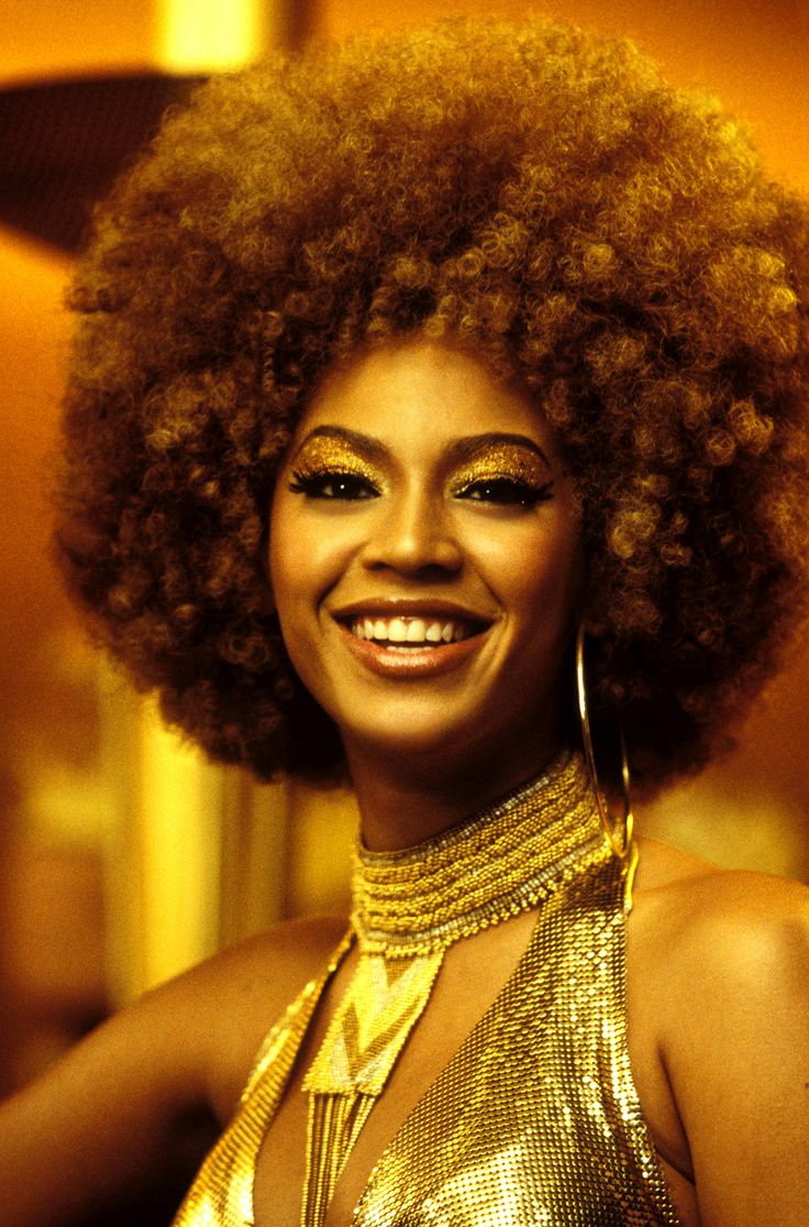 a woman with an afro smiling and wearing gold necklaces on her neck, standing in front of a multi - colored background