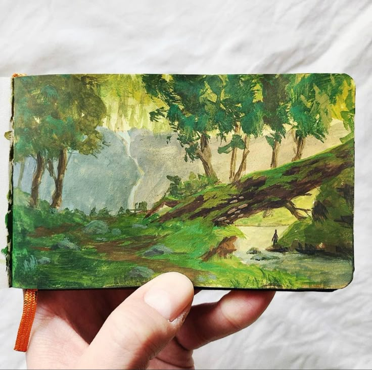 a hand holding an open book with a painting on it's cover and trees in the background