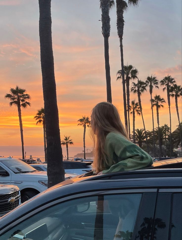 a woman sitting on the roof of a car looking out at palm trees and sunset