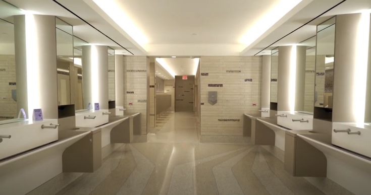 an empty public restroom with sinks and mirrors on the wall, lights on either side of the stalls