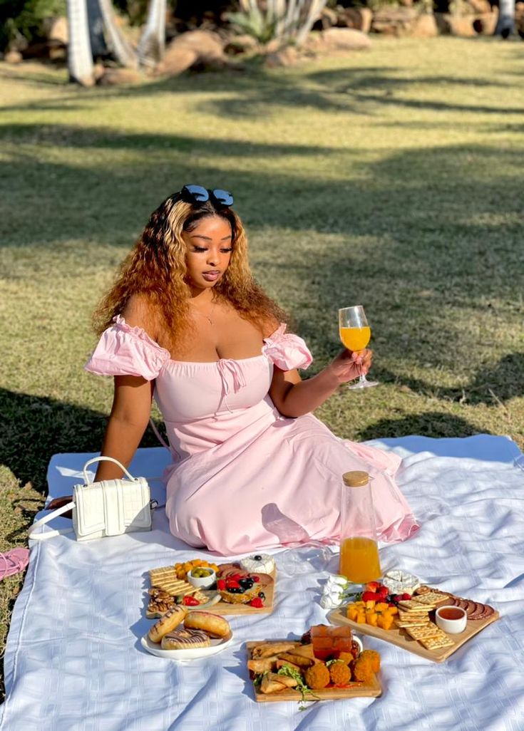 a woman in a pink dress is sitting on a blanket with food and wine glasses