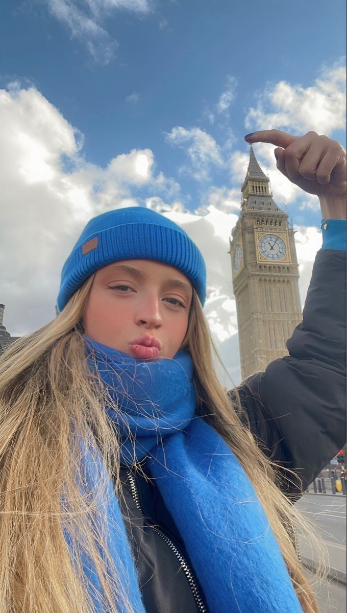 a girl with long blonde hair wearing a blue hat and scarf in front of the big ben clock tower