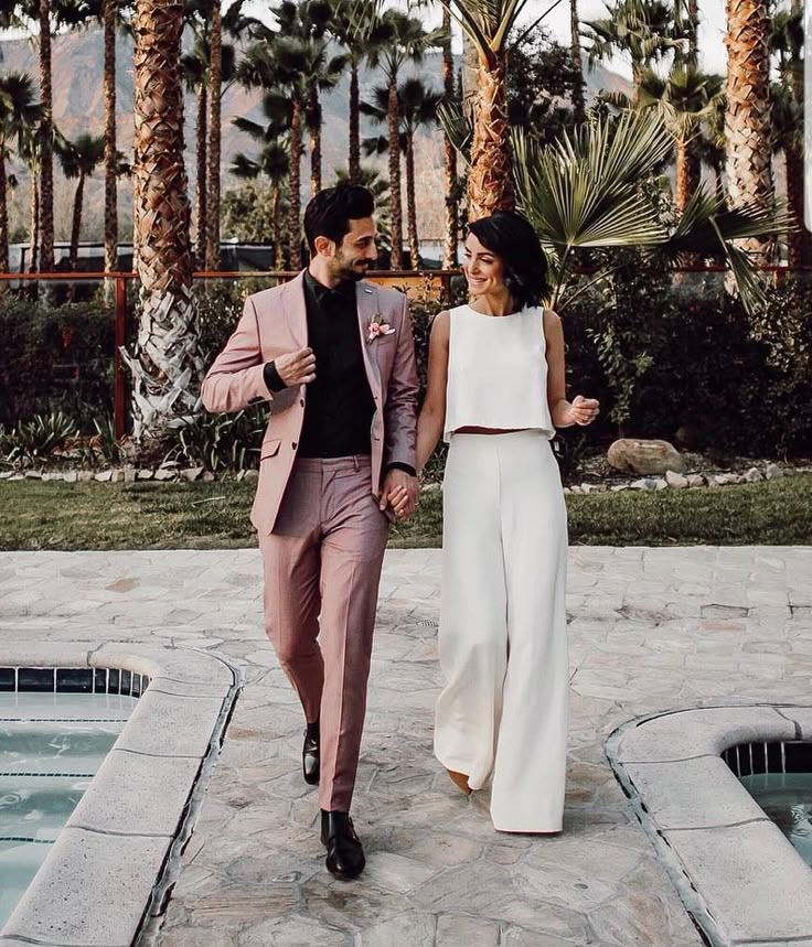 a man and woman holding hands walking next to a pool with palm trees in the background