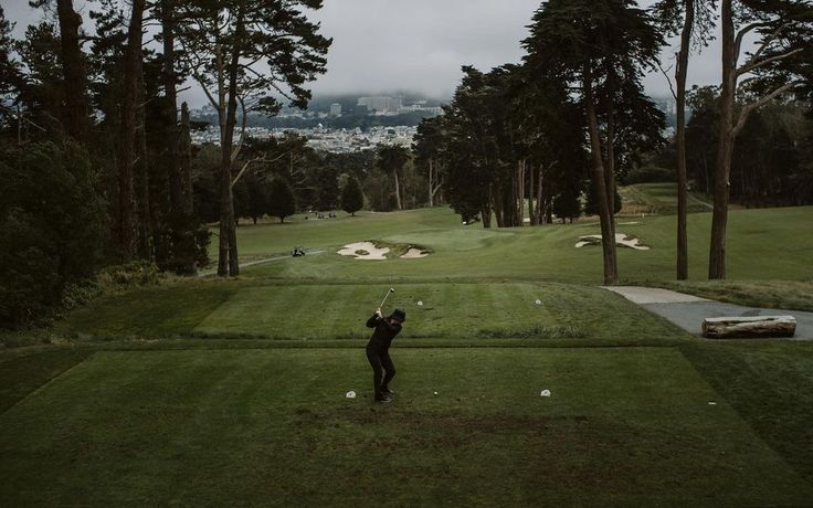 a man swinging his golf club at the green in front of some trees on a cloudy day