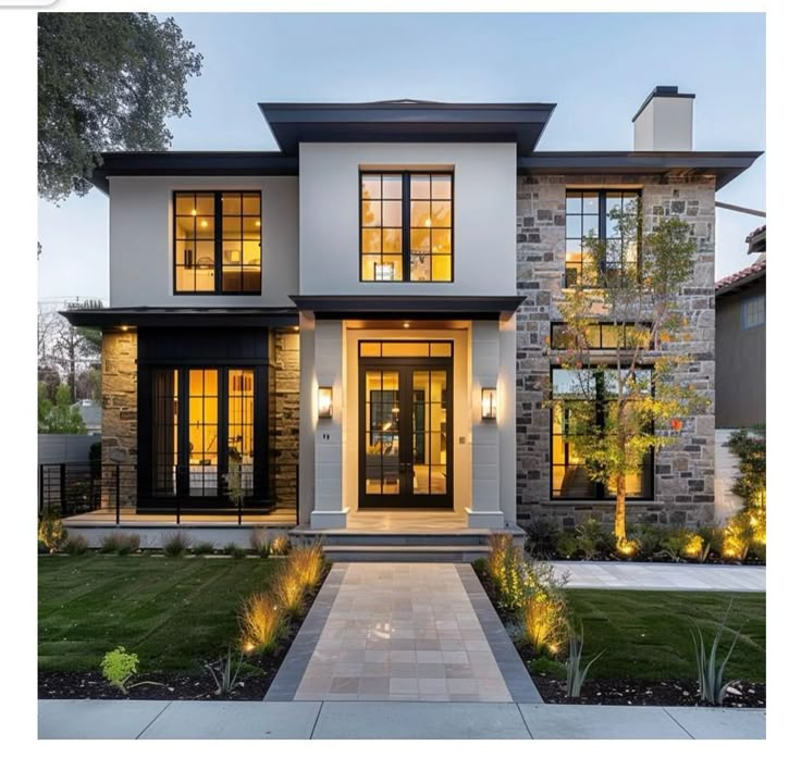 a modern house with lots of windows and lights on the front door, along with landscaping