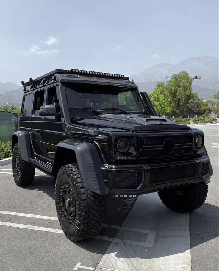 a black mercedes g - class is parked in a parking lot with mountains in the background