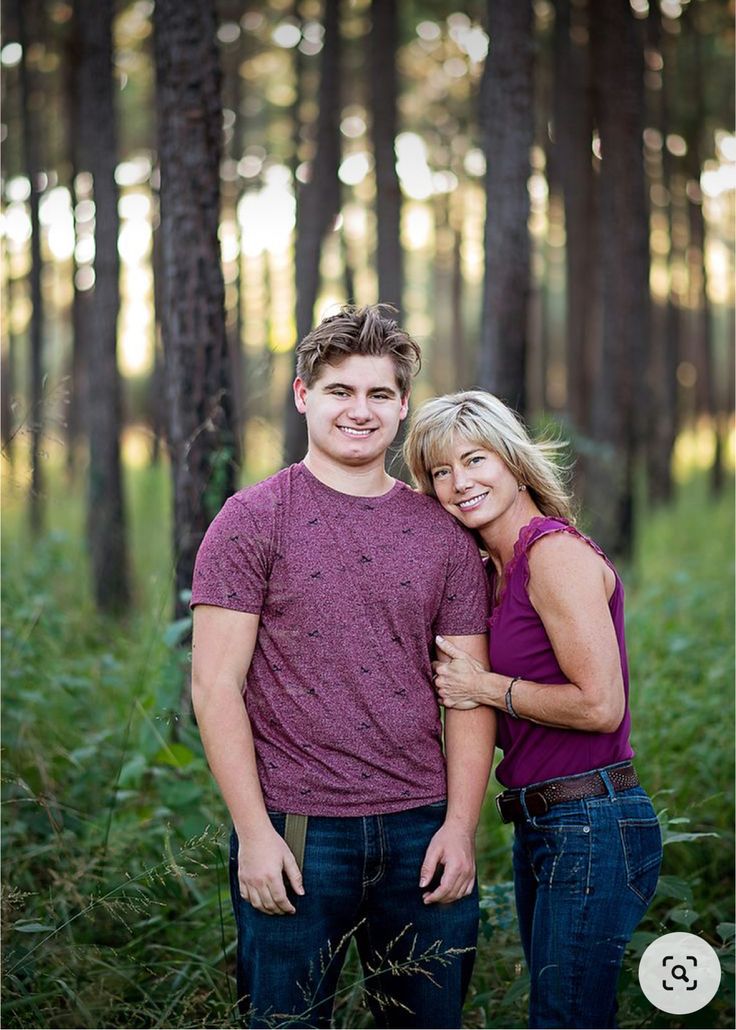 a man and woman standing next to each other in front of tall grass with trees behind them