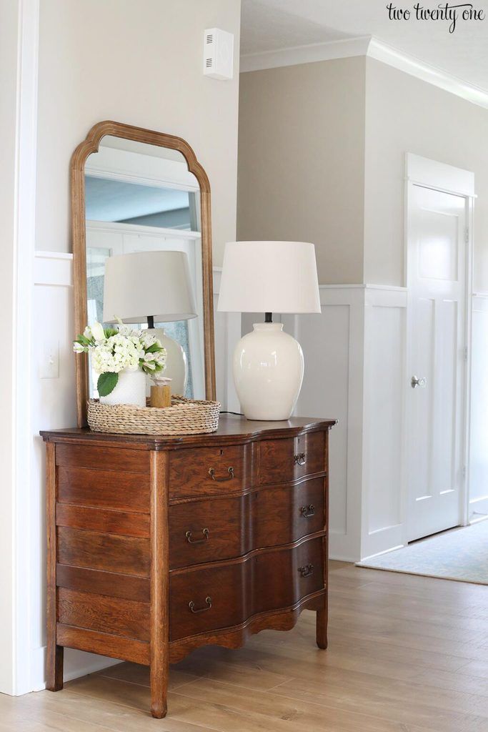 a wooden dresser topped with a white vase filled with flowers next to a large mirror