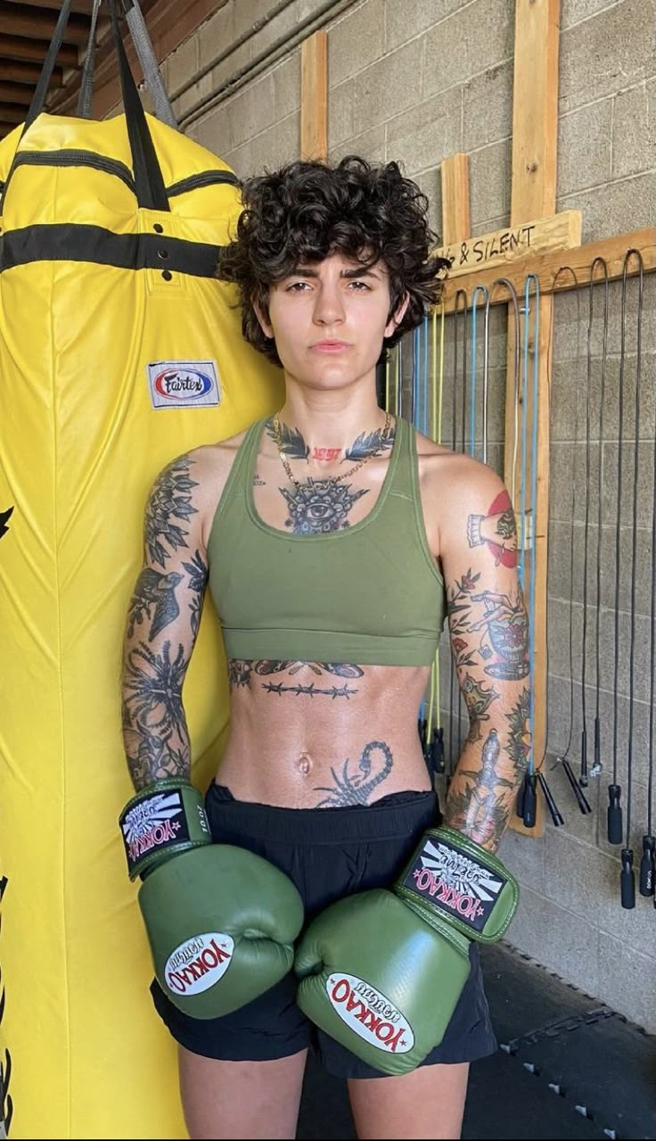 a woman with tattoos standing in front of a surfboard and holding two green boxing gloves