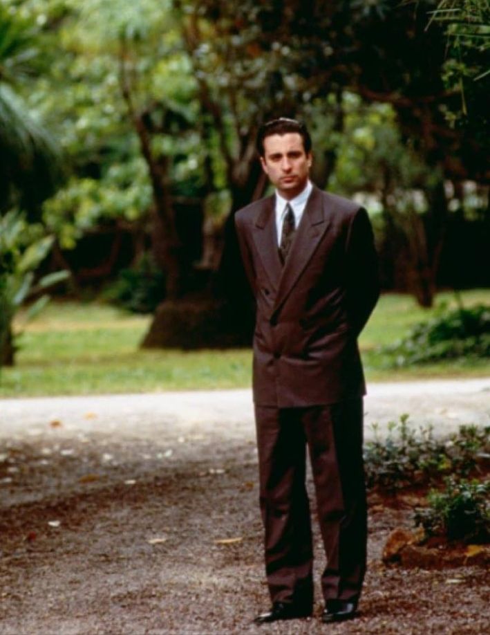 a man in a suit and tie standing on a dirt road with trees behind him