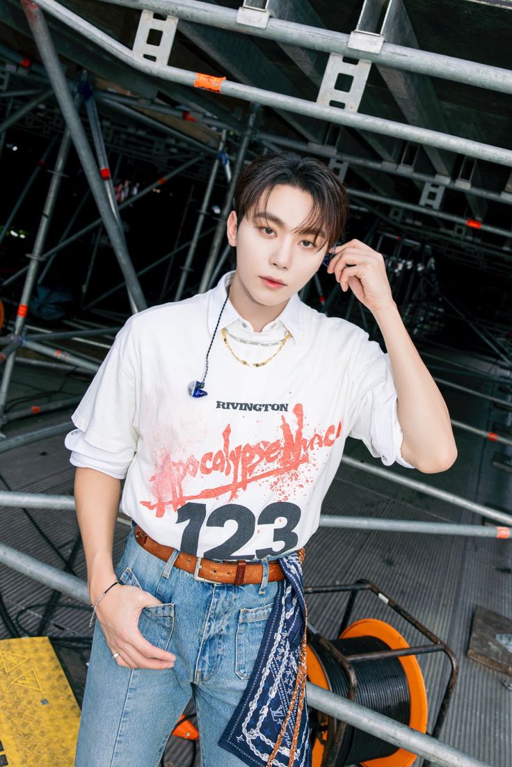 a young man standing in front of a metal structure wearing a white shirt and blue jeans