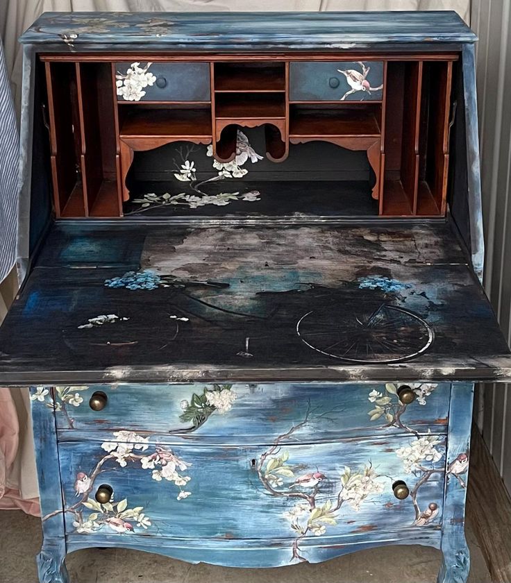 an old desk painted blue with flowers and birds on the top, sitting in front of a wall