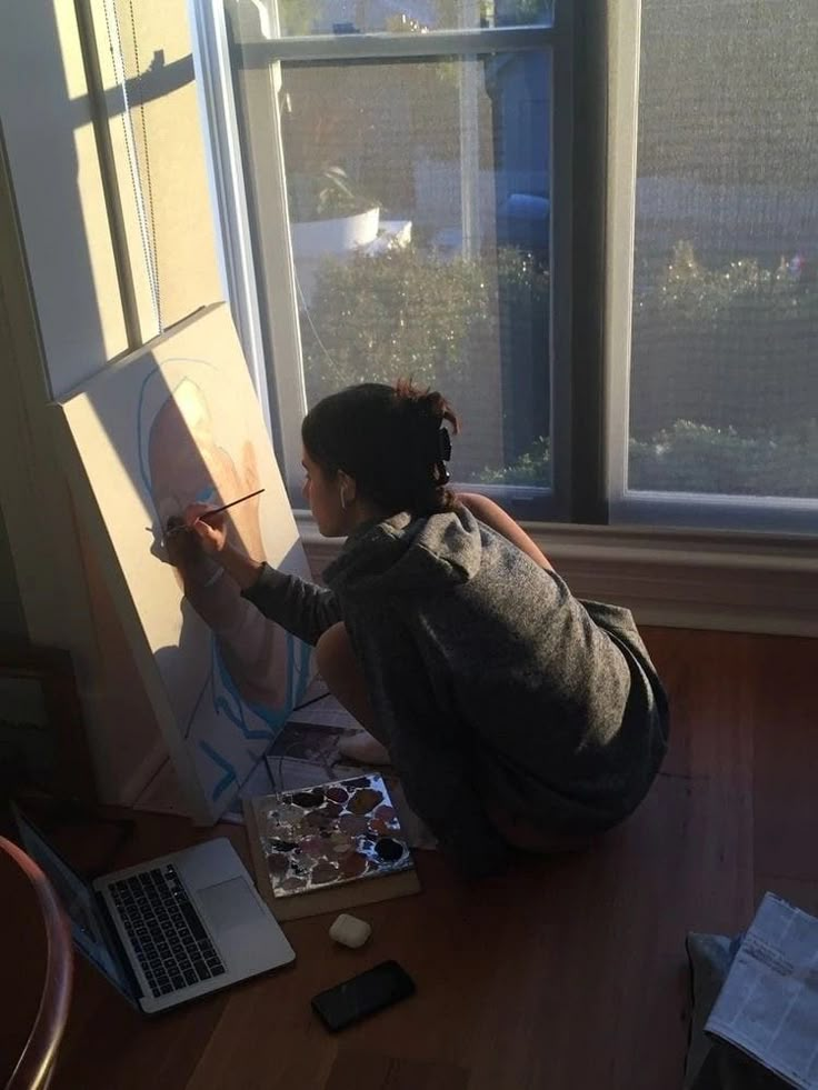 a woman is sitting on the floor and painting with her laptop in front of her