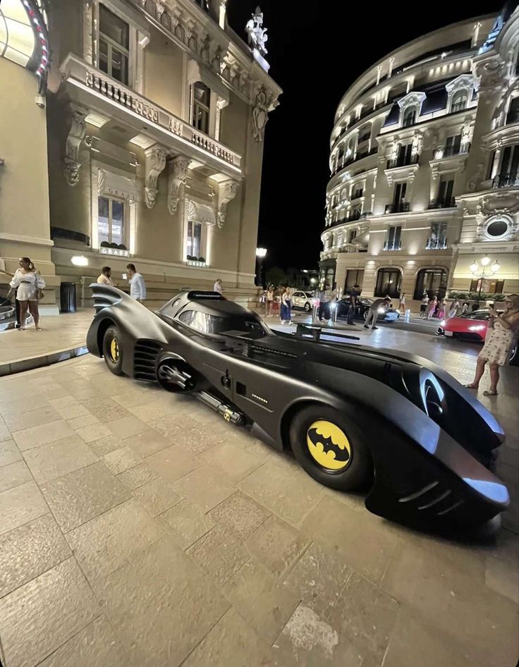 a batman car is parked in front of a building at night with people walking around