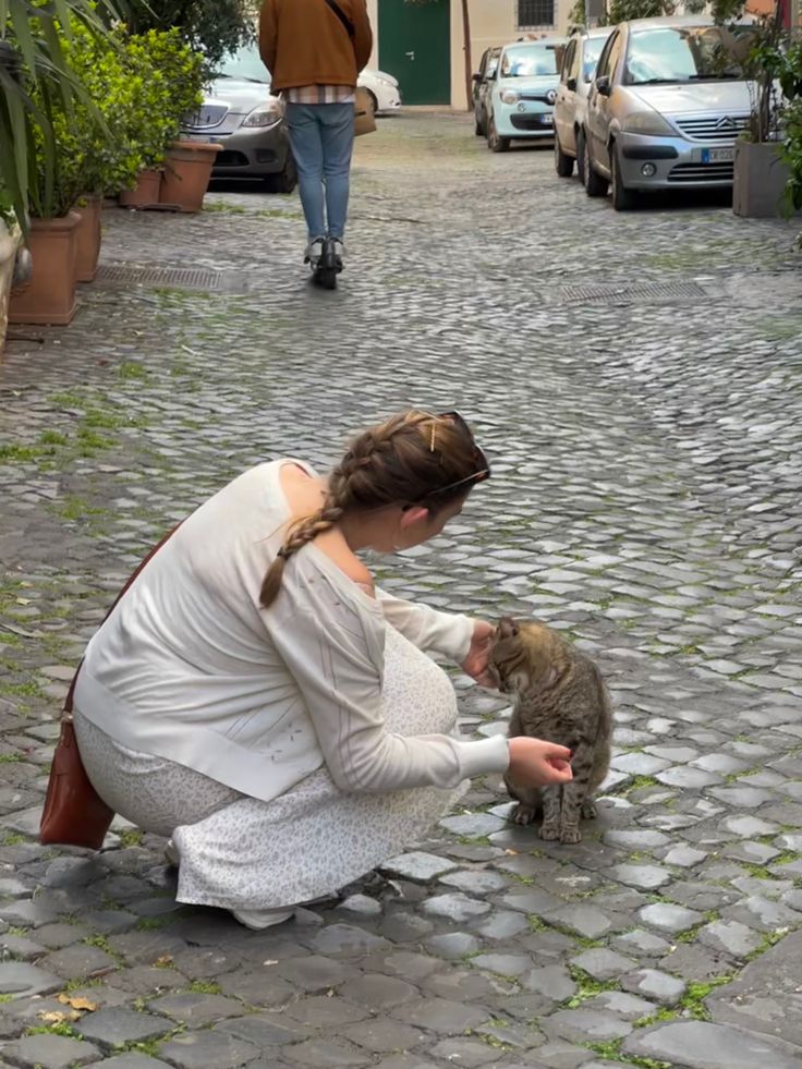 a woman kneeling down petting a cat on top of a cobblestone street