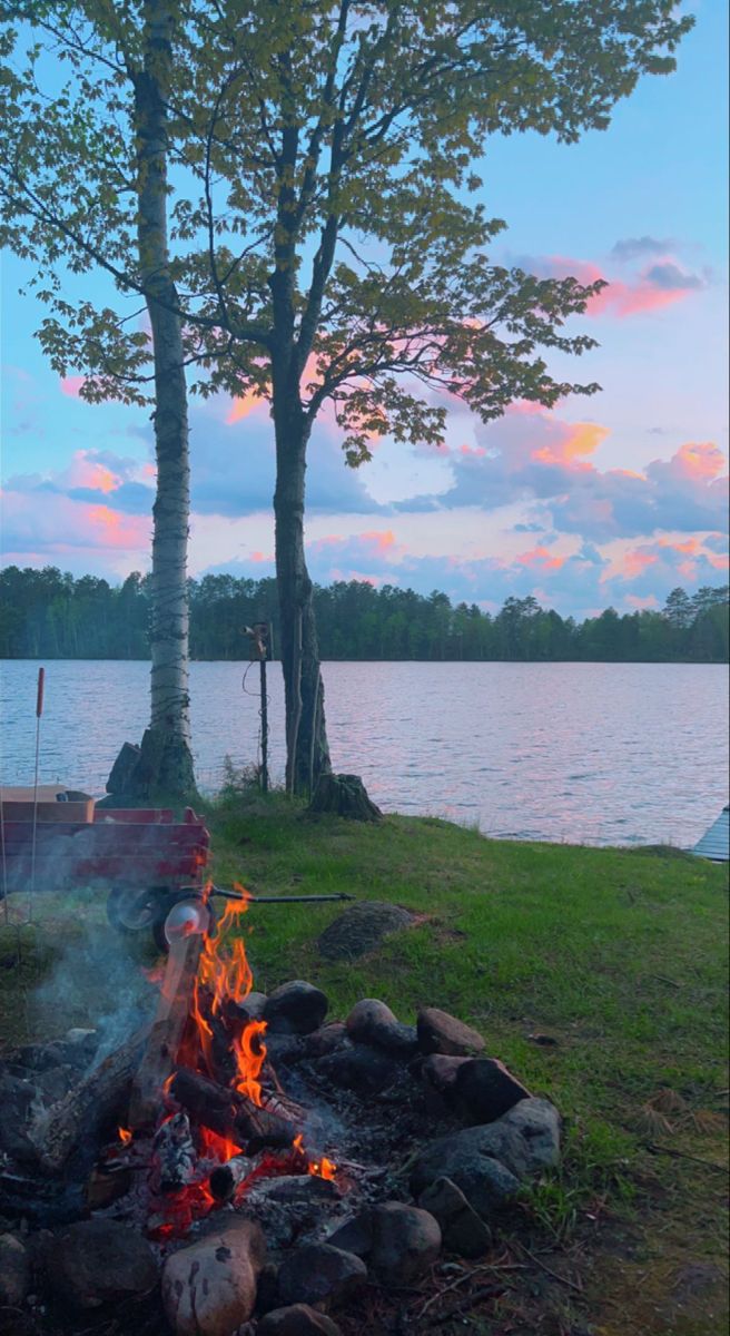 a campfire next to a lake with trees and water in the background at sunset