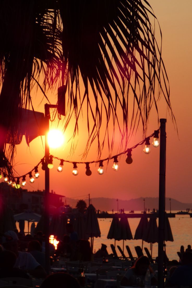 the sun is setting over an ocean with people sitting at tables and umbrellas in the foreground