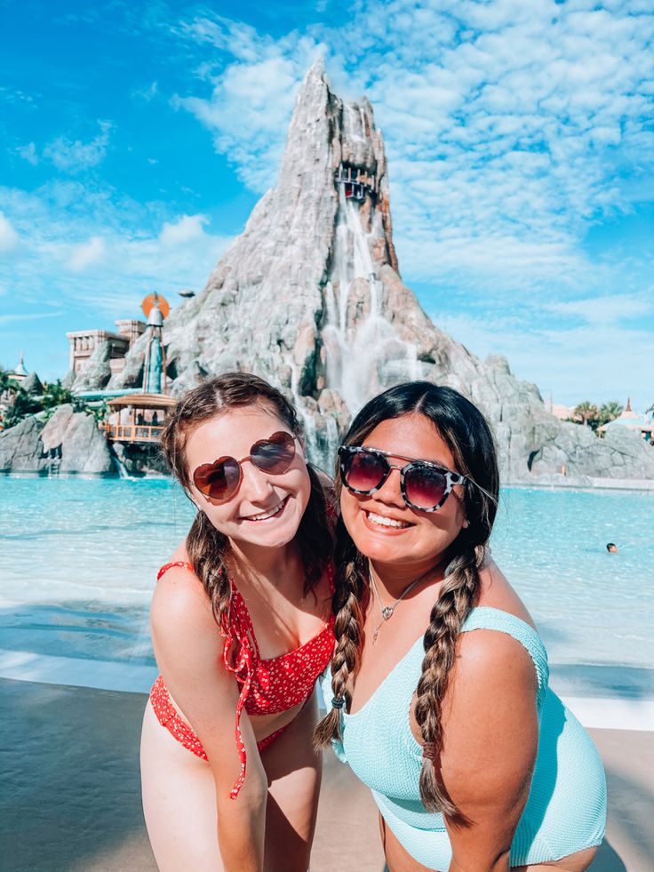 two women in bathing suits pose for a photo on the beach with a mountain behind them